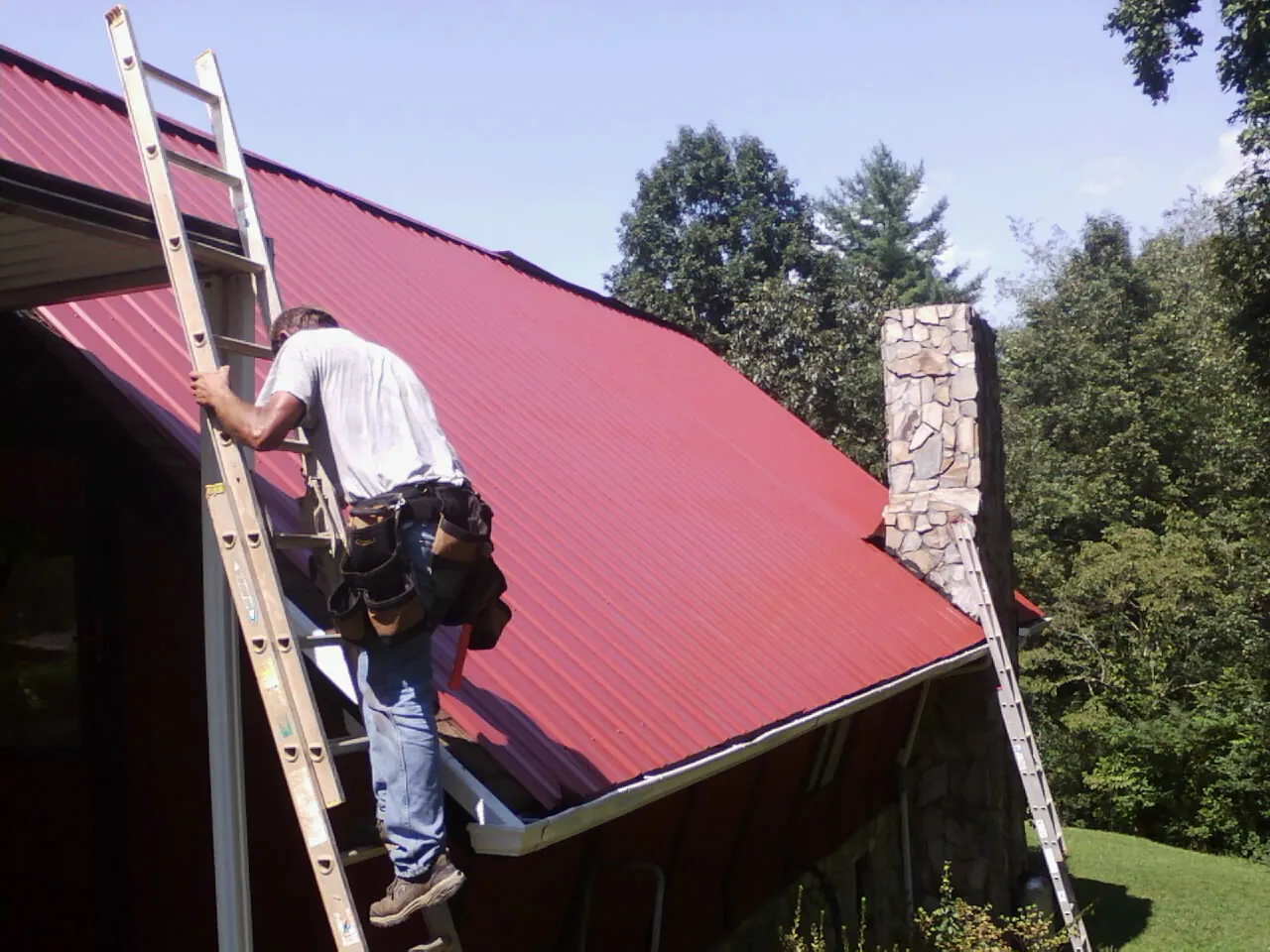 Roofer on a ladder beside a red metal roof and stone chimney — Soffit & Fascia Repair by Roofing Brothers in Rancho Santa Margarita